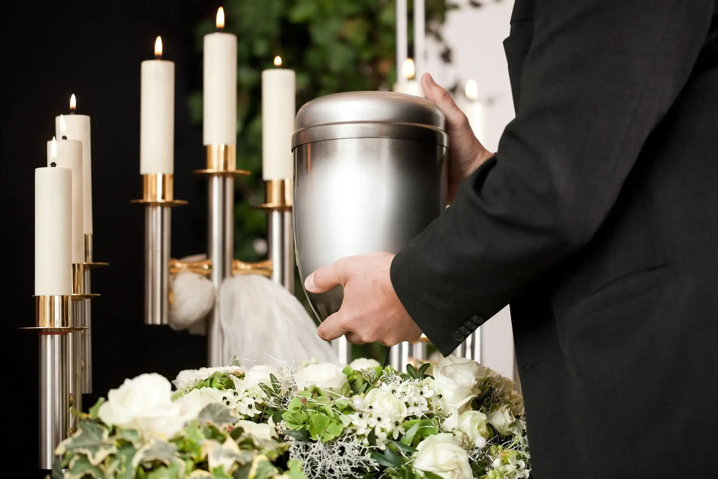 Urnă funerară pentru cenușă, ținută de un angajat al casei funerare, între lumânări și flori albe